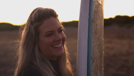 Mujer-Vistiendo-Traje-De-Neopreno-Sosteniendo-Una-Tabla-De-Surf-Disfrutando-De-Unas-Vacaciones-De-Surf-En-La-Playa-Mientras-Se-Pone-El-Sol