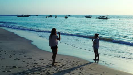 Mujer-Turista-Tomando-Una-Foto-De-Un-Amigo-En-La-Playa-De-La-Puesta-De-Sol-Que-Está-Rodeada-De-Un-Océano-Azul-Brillante-Y-Veleros---Plano-General