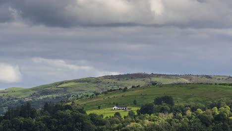 Zeitraffer-Der-Ländlichen-Landwirtschaftslandschaft-Mit-Einem-Bauernhaus-Auf-Einem-Hügel-Mit-Wald-Im-Vordergrund-An-Einem-Bewölkten-Sommertag-In-Irland