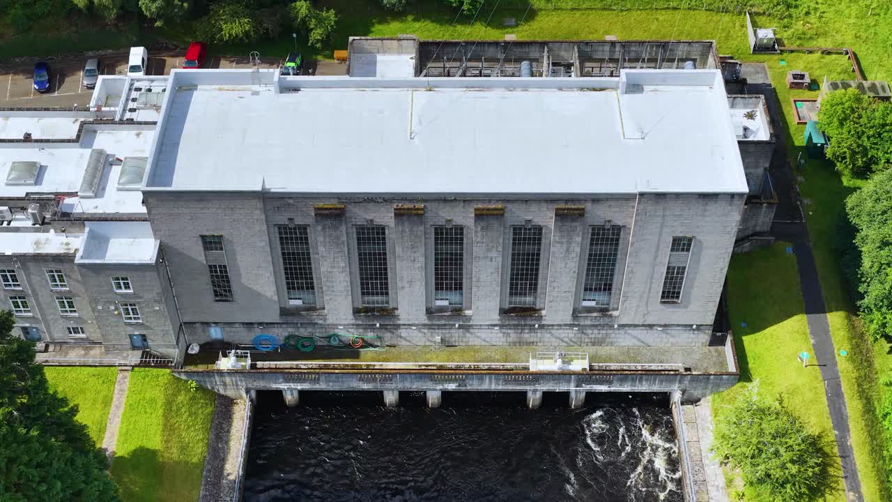 Drone slowly ascends above hydroelectric power station, river, and green landscape in bright daylight