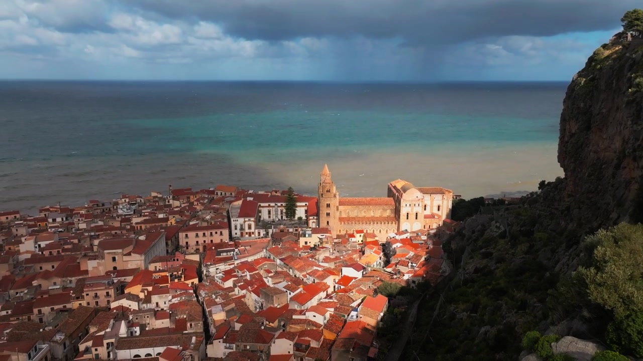 Cefalù: Ripresa aerea con drone in tilt-up di una falesia rocciosa di montagna che rivela una storica città costiera antica e il Mar Mediterraneo, Sicilia Italia