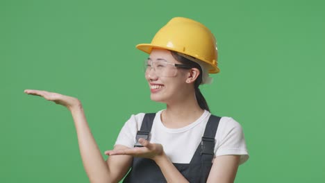 close up of asian woman worker wearing goggles and safety helmet smiling and pointing to side while standing in the green screen background studio