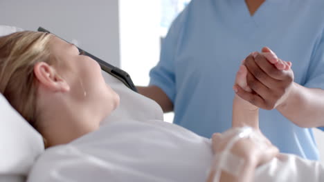 diverse female doctor and girl patient lying on bed holding hands in hospital, slow motion