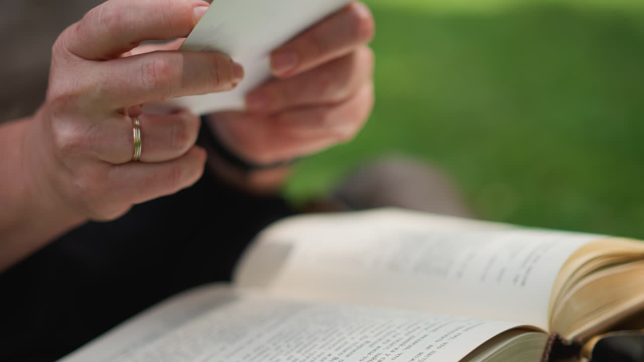 Close up of lady holding open book with both hands, turning to next page under natural light, delicate focus showing calm emotion and nostalgia as she pauses over photo, surrounded by blurred green