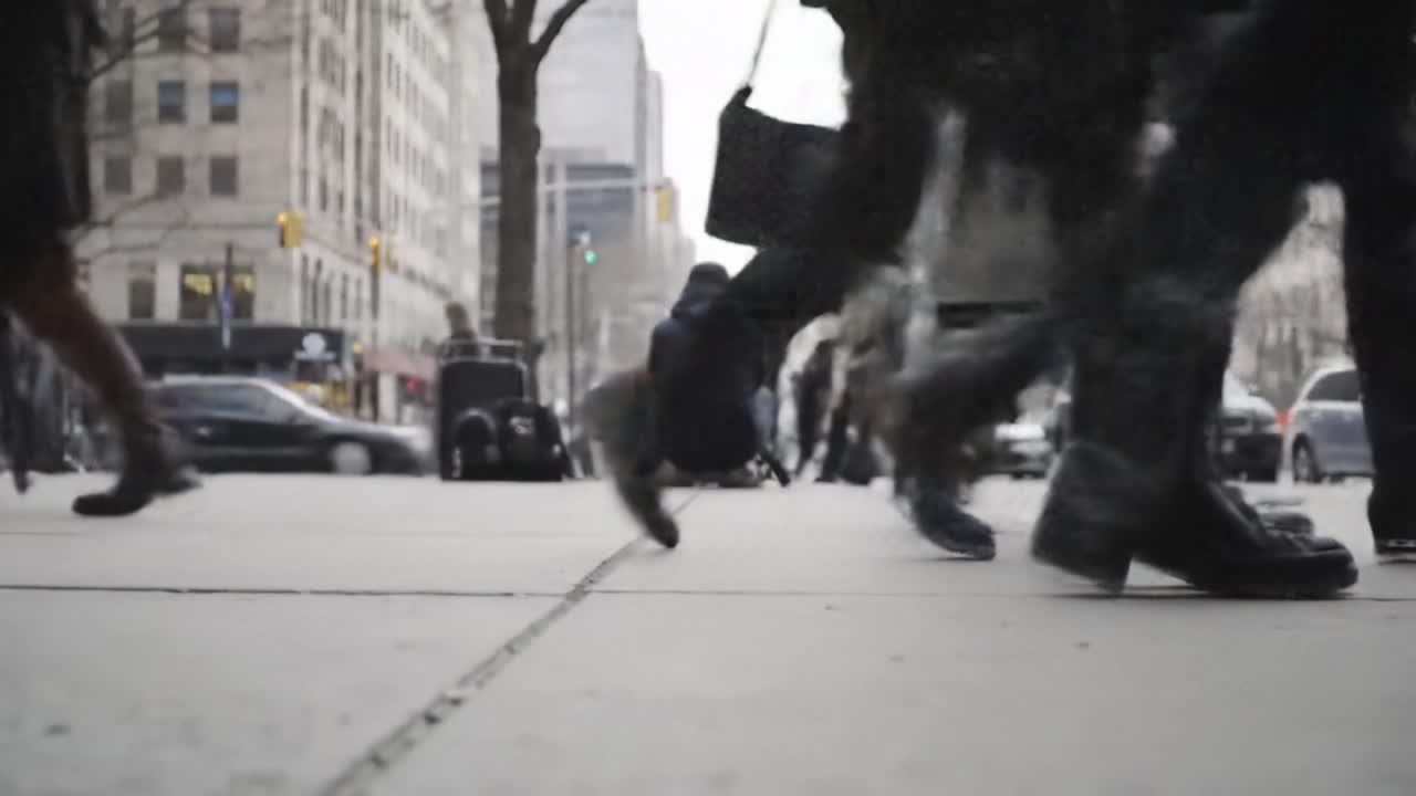 People hurry along the sidewalk in a bustling urban environment, their shoes striking the pavement amid the sounds of the city. The scene captures the energy of daily life in a metropolitan area.