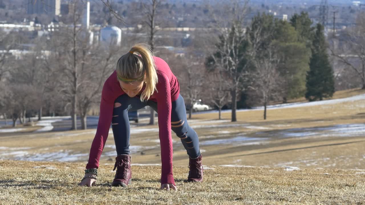 mujer rubia con botas y ropa deportiva estira las piernas en el parque sobre la hierba amarilla en invierno