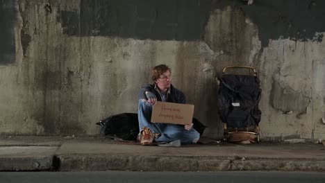 Wide-Shot-of-a-Disheveled-Homeless-Young-Adult-Male-Holding-an-Anything-Helps-Sign-in-Shame