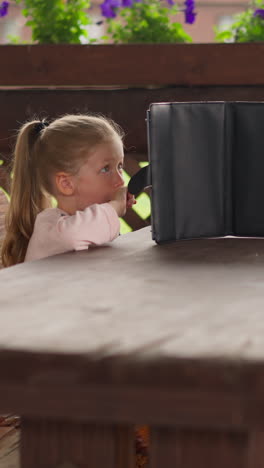 adorable little girl with tablet computer sits and waits for rain ending at empty wooden table in restaurant at eco hotel on stormy day slow motion