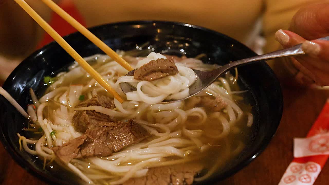 A person savors a bowl of Vietnamese pho with chopsticks and spoon in a cozy setting