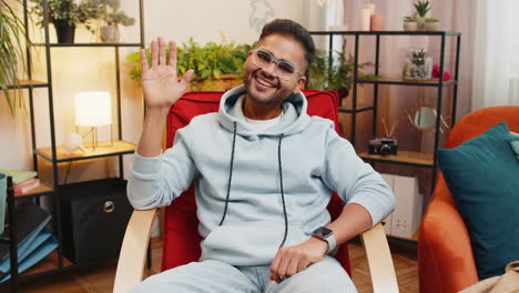 a young man in a blue hoodie and glasses waves and smiles while sitting in a chair in a living room.