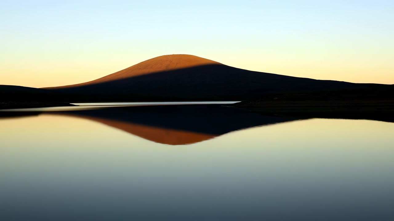 Solitary rounded hill stands over glassy lake, while warm light fades into deeper silhouette, reflection darkens, and broad shadow moves across midsection, creating serene natural symmetry and quiet