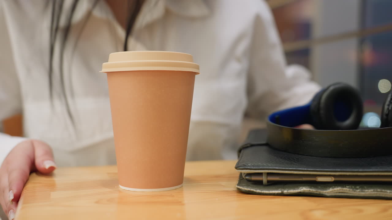 Close up of woman gently placing coffee cup on wooden table with headphones and tablet nearby, as soft bokeh lights of moving cars glow outside through large glass panel in cozy indoor setting
