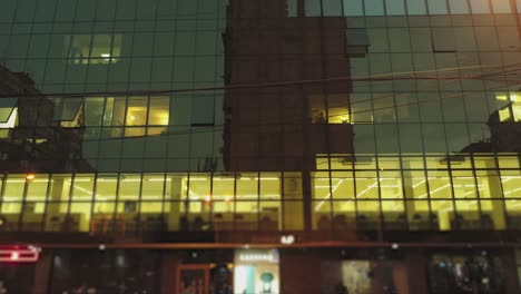 4k outside view of business district building , mall with wall mirror windows at evening or night .  reflection of city , people , cars in mirrored walls . drone camera moving . people working inside