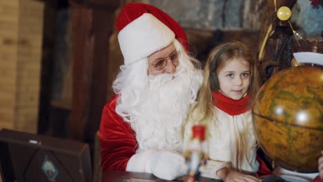 Pretty little girl sitting with Santa Claus in front of the Globe model. Santa in red and white suit looking at globe together with a child at Christmas.
