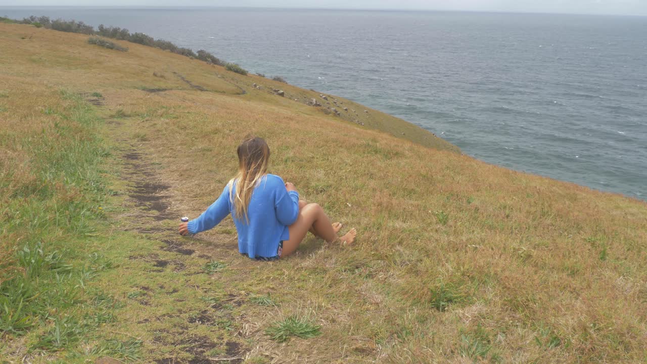 통조림 음료를 즐기고 초승달 머리 전망대에서 차가운 바닷 바람을 느끼면서 풀밭에 앉아있는 소녀 - 호주 뉴 사우스 웨일즈의 큰 노비 반도