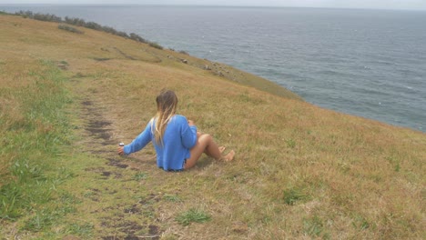 girl sitting on grass while enjoying canned drink and feeling cold ocean breeze at crescent head lookout - big nobby peninsula in nsw, australia