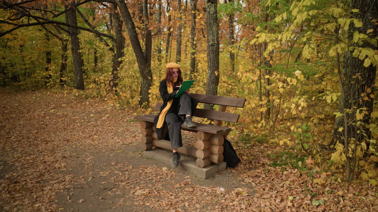 평온한 환경에서 나무로 된 공원 벤치에 앉아, 한 다리가 벤치 위에, 다른 다리가 바닥에, 바닥에 가방을 가지고, 황금 가을 잎으로 둘러싸인 평화로운 자연에서 스케치에 깊이 집중 한 예술가.