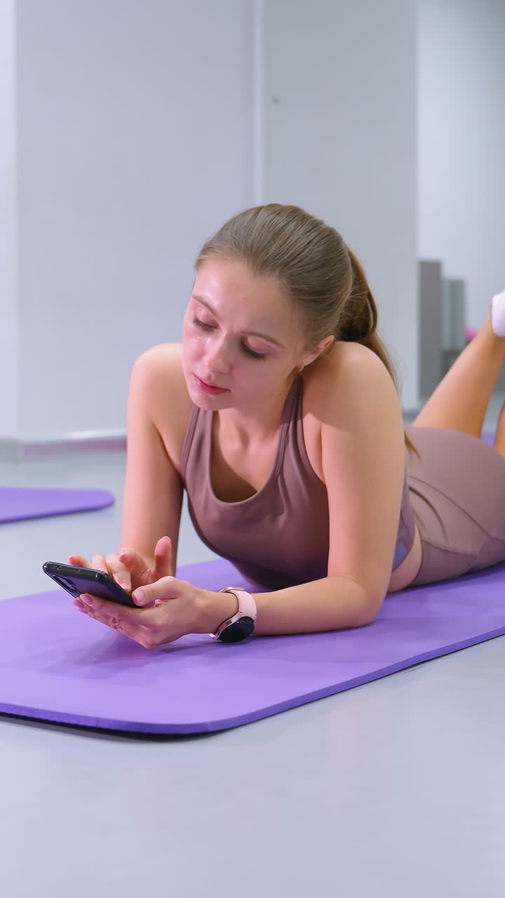 mujer joven yace en la alfombra de yoga, centrado en el teléfono, levantando las piernas ligeramente, el fondo cuenta con grandes espejos y gabinetes de almacenamiento en un estudio de fitness moderno