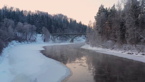 Bosque-Cubierto-De-Nieve-Y-Río-Helado-Con-Puente-Al-Amanecer,-Disparo-De-Drones