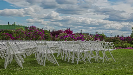 static shot of rows of wooden folding white chairs on the green grass garden floor with white clouds passing by on a cloudy day in timeplase