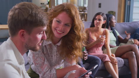 young couple sitting on sofa at home talking and looking at mobile phone together