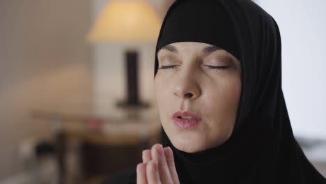 close-up portrait of attractive muslim woman holding hands together and praying. lady in black traditional hijab talking with god and closing eyes. camera moving around from left to right.