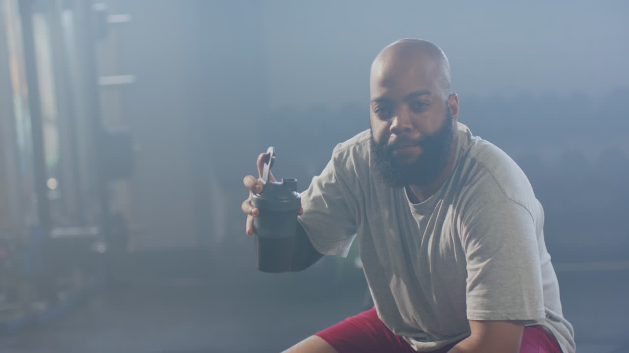 Uomo afroamericano su una panca da palestra si reidrata da uno shaker dopo una serie, mostrando il tappo e sorridendo.