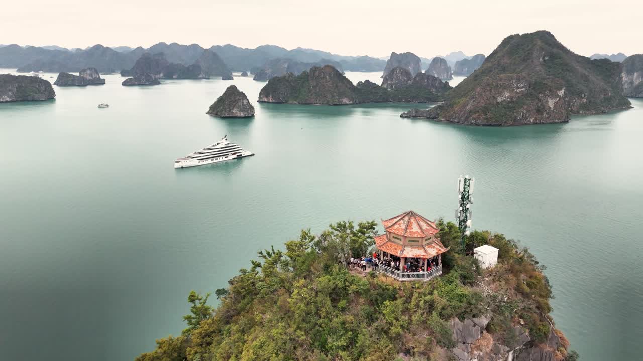 A dramatic aerial view of Ha Long Bay, Vietnam, featuring a cliffside pavilion, lush greenery, limestone islands, and a luxury yacht gliding across calm emerald waters under an overcast sky