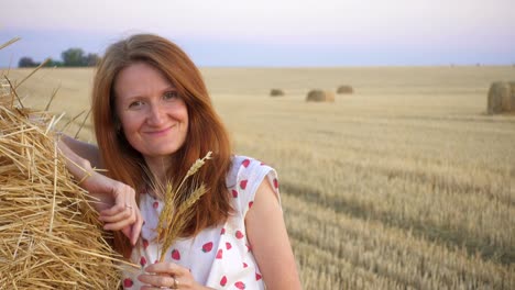 outdoor evening portrait of a beautiful young smiling girl near the round bale of hay in a wheat field