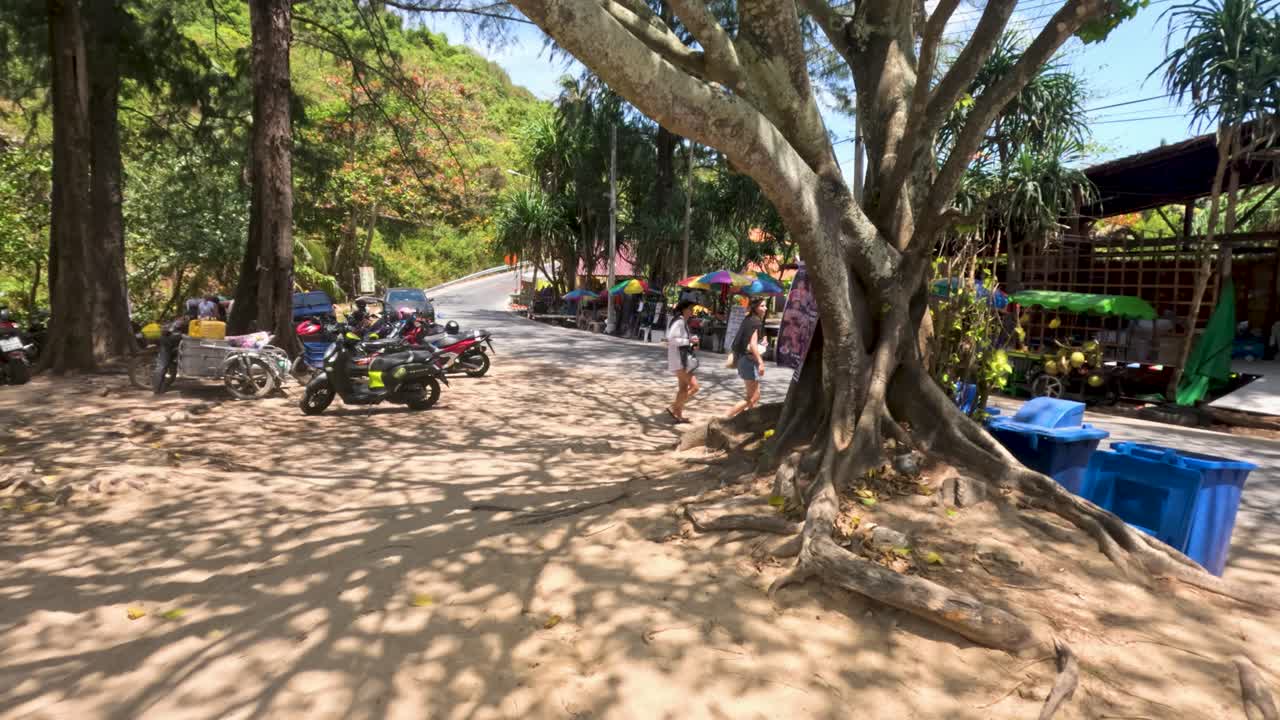 Moped riders travel past a jungle cafe in Phuket, Thailand. Sunlight filters through trees, creating a vibrant, tropical atmosphere