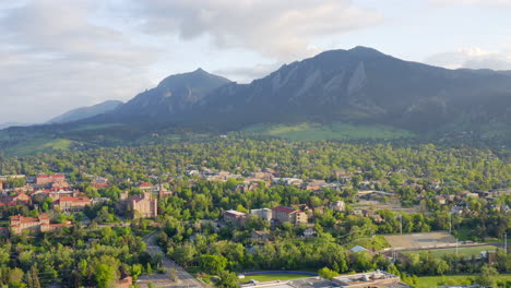 Aerial-pan-left-of-beautiful-flatiron-mountain-vista,-bright-green-trees,-and-CU-Boulder-campus-in-Boulder-Colorado-during-an-evening-sunset-with-warm-light-on-the-summer-landscape