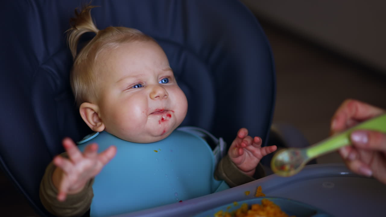 Unrecognized mom feeding her little baby from spoon. Kid makes faces as he eats the food. Close up.