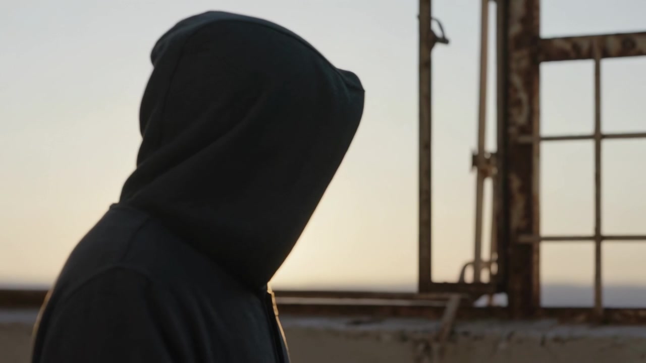 Gående hettekledd skikkelse beveger seg fremover over et tak i solnedgang etter en pause, og nærmer seg et rustent gitter