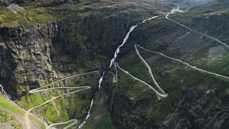 vehicles travel along the winding trollstigen road, with a waterfall crashing down the mountain beside the road. a tilt-shift video. aerial view.