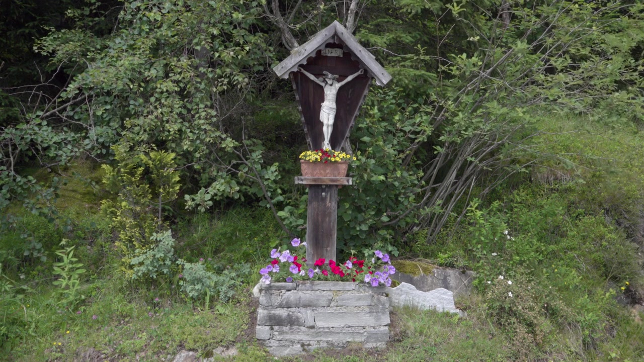 A wooden Christian cross surrounded by flowers, bushes and grass in the open air