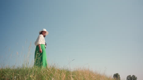 woman in wide-brimmed hat and green skirt stands near grassy cliff edge, holding camera while looking out into open horizon under bright clear sky, surrounded by tall grass