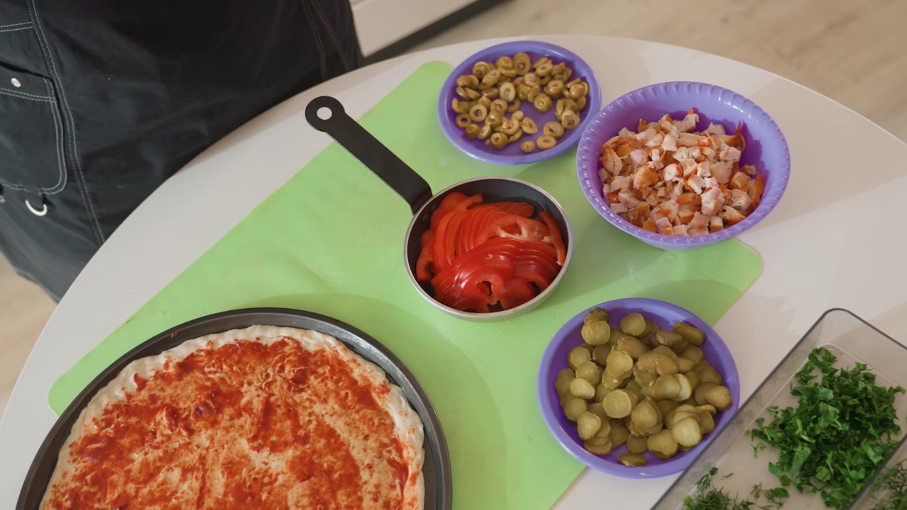 Vista de cima de pessoa montando pizza fresca em cozinha caseira casual, molho circular se espalhando sobre massa crua enquanto colher, tigela, presunto picado e ervas estão próximos durante o processo de cozimento