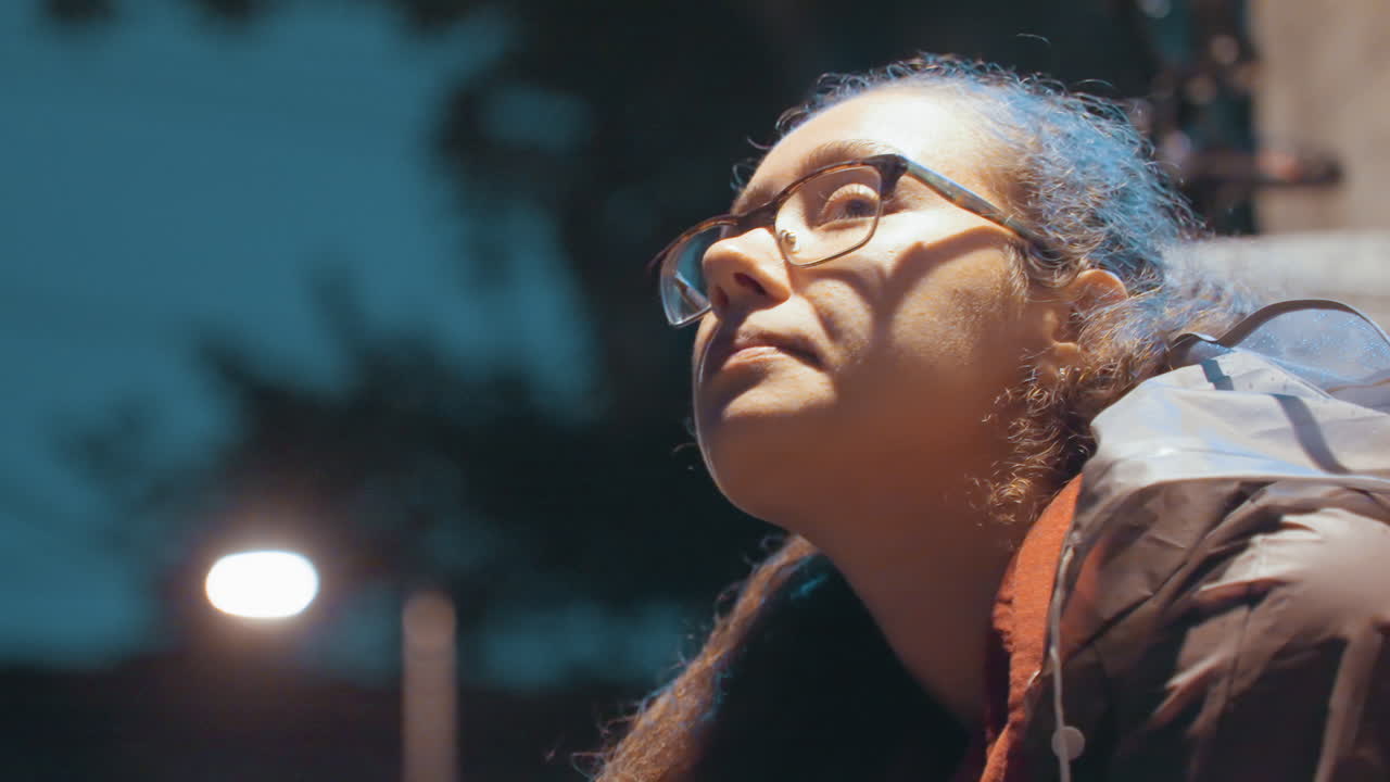 Young woman with snack looks upward. Portrait of woman holding snack under lamplight in city night. Nighttime city street capturing young woman gazing upward with contemplative expression