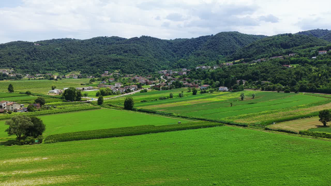 田園風景の中に広がる緑豊かなパッチワーク状の畑の空撮。背景には村と森林に覆われた丘陵地帯。農業や旅行コンテンツ、イタリア、ベリチ丘陵。