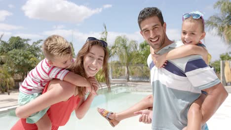 Portrait-of-happy-caucasian-parents-carrying-children-at-swimming-pool-at-beach-house