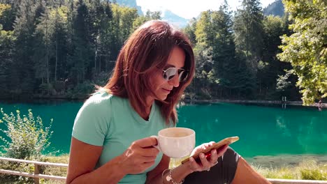 mujer mayor deportiva y atractiva relajándose junto a un lago en la naturaleza después de hacer ejercicio en verano, por teléfono, tomando café y echando un vistazo rápido a la cámara