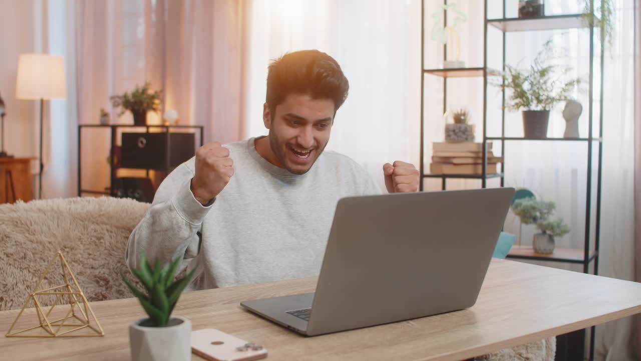 Young indian man using laptop celebrating after receiving important email and good news at table
