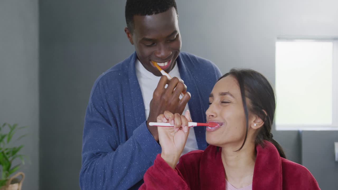 Video of happy diverse couple brushing teeth and smiling in bathroom at home