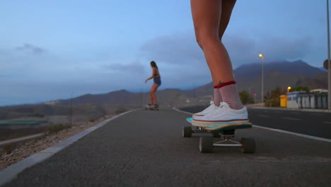 Against-the-backdrop-of-the-setting-sun,-a-beautiful,-stylish-skateboarder-rides-in-shorts-on-a-mountain-road,-revealing-the-mountains'-stunning-scenery-in-slow-motion