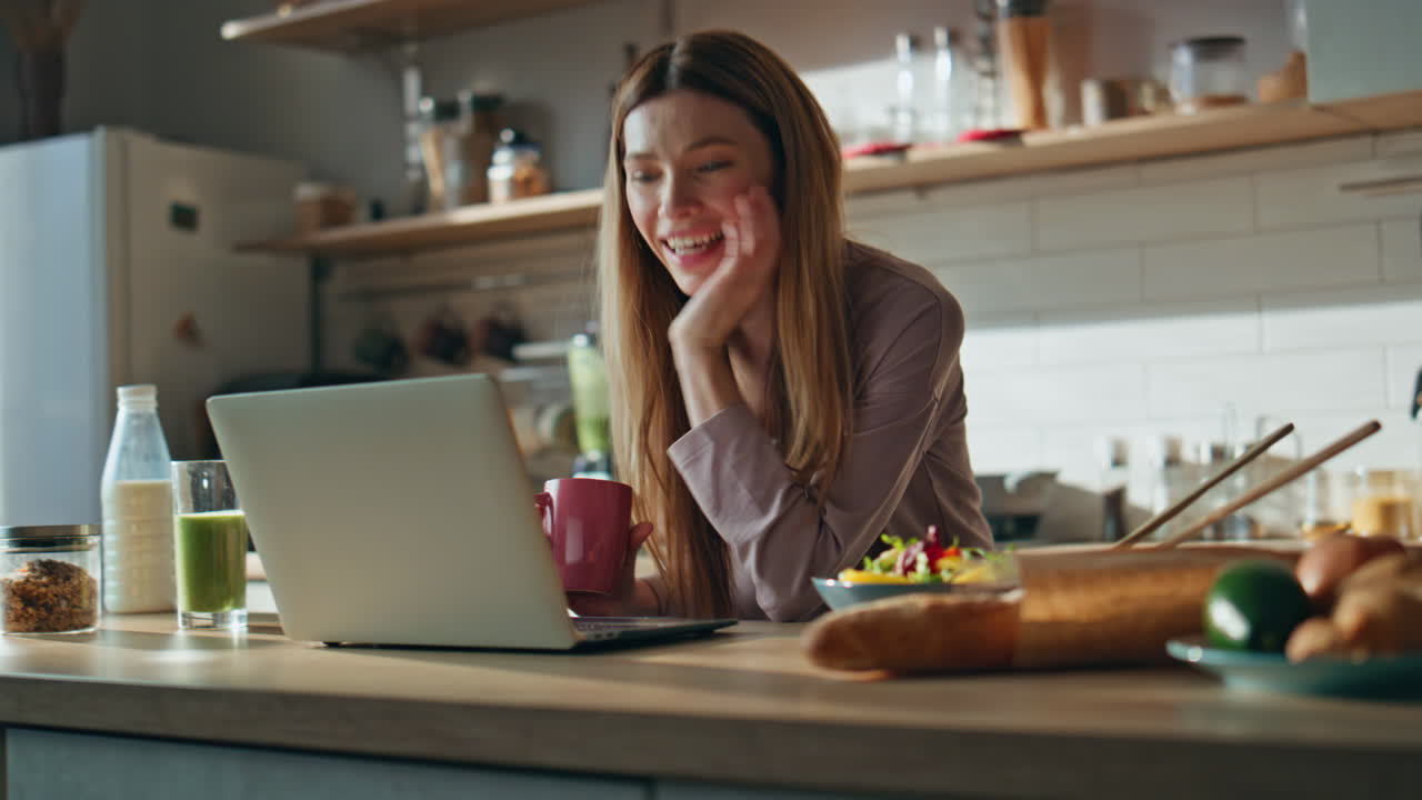 Smiling housewife videocalling laptop holding coffee cup at cozy kitchen closeup