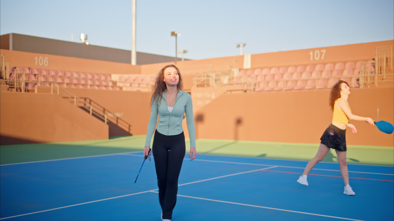 Two women with curly hair playing pickleball on a blue court on a sunny day