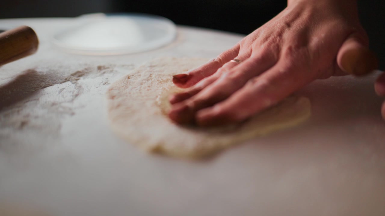 Una mano preme l'impasto su una superficie infarinata. Il centro oleoso luccica sotto le dita. La calda luce della cucina evidenzia la consistenza. Perfetto per progetti di cucina, culturali o legati al cibo.
