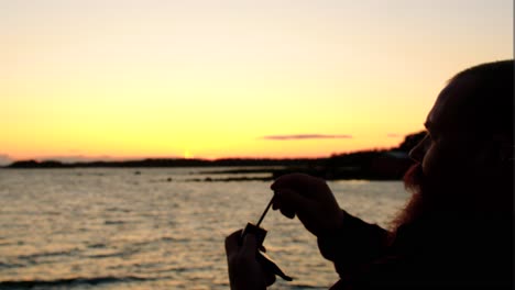 adult male with short hair and long moustache beard sitting by the sea preparing his pipe filled with tobacco for a smoke with a posh tool accessory wonderful golden hour sunset independent meaningful