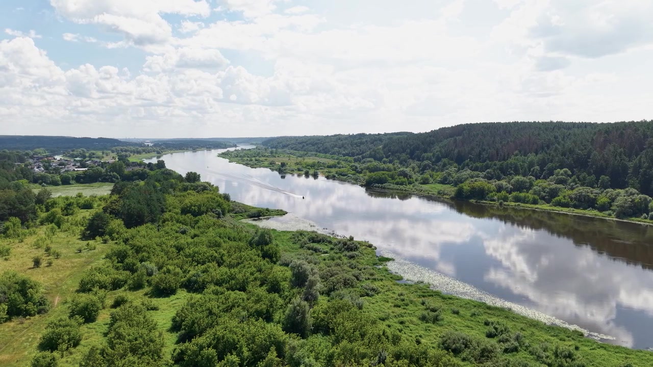 Riprese aeree mostrano un ampio fiume che serpeggia tra foreste verdi e campi aperti sotto la luce del sole splendente, riflettendo il cielo blu e le nuvole. Paesaggio naturale pacifico e incontaminato, nessuno visibile.