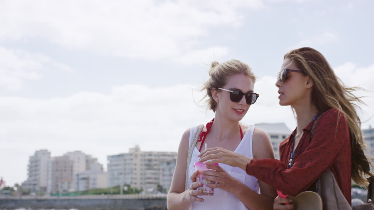 두 소녀 여자 친구가 술을 나누고 해변 마을의 산책로를  ⁇ 고 편안한 휴가를 즐기고 있습니다.